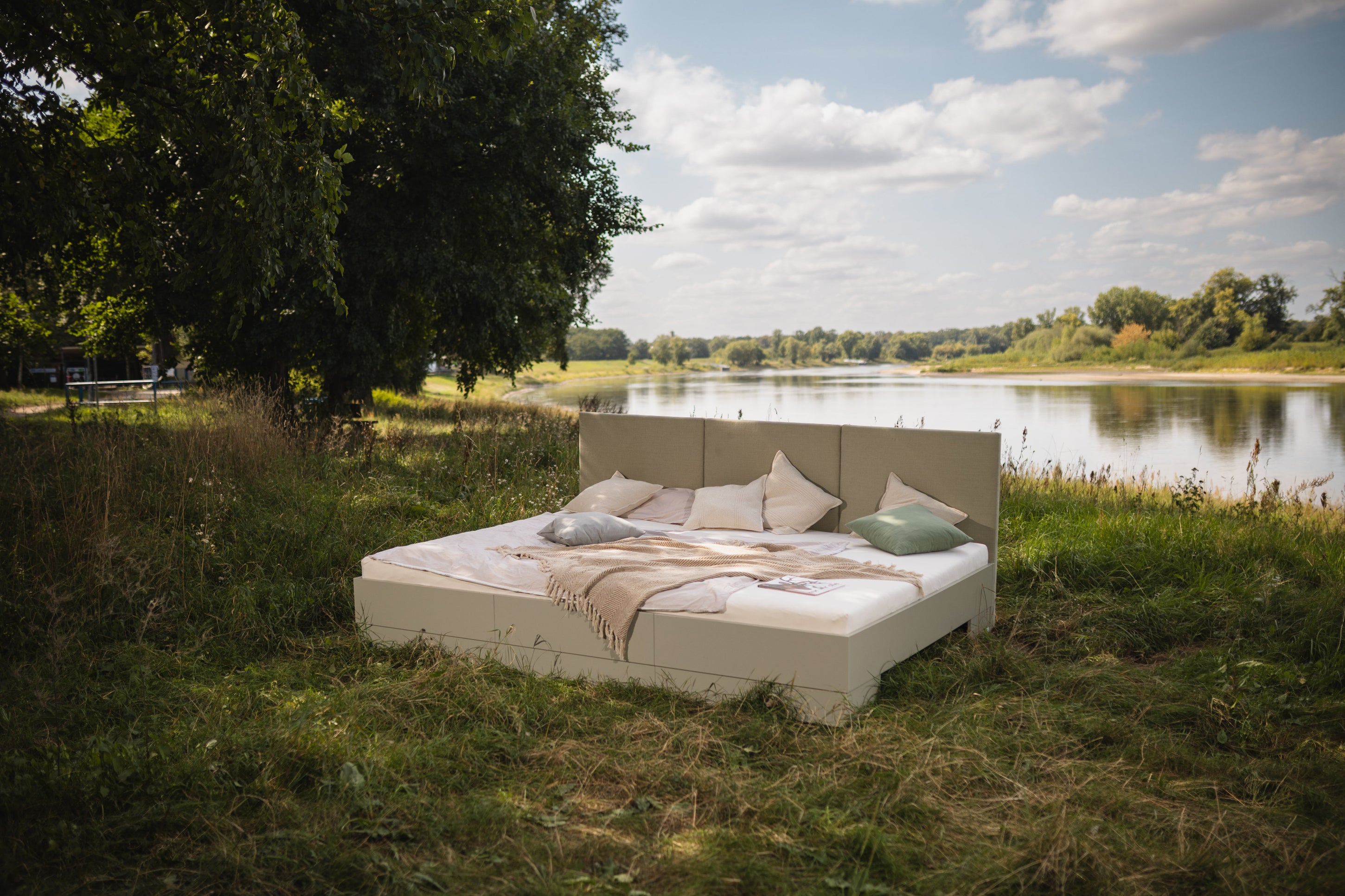 Familienbett Magnolia mit Kopfteil aus Stoff in Salbeigrün steht auf einer Wiese an der Elbe