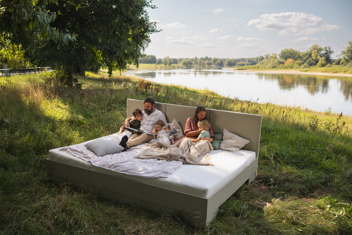 Familienbett Magnolia in Salbeigrün steht auf einer Wiese an der Elbe unter einem Baum. Die Familie sitzt auf dem Bett und liest. 